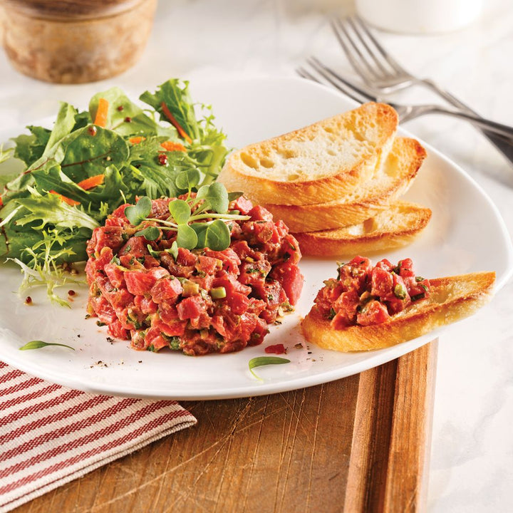 Tartare de bœuf présenté dans une assiette avec croûtons, cornichons et salade, accompagné de sachets de sauce à tartare Aliments DKL.